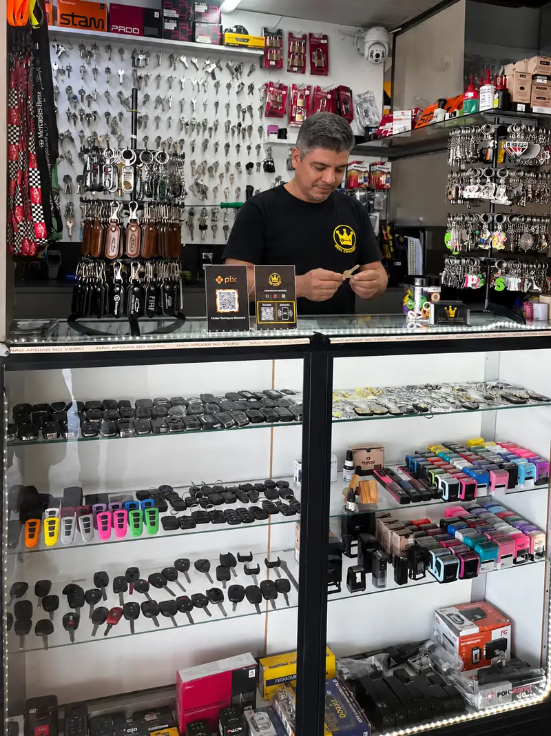 Cleber no balcão do Real Chaveiro olhando de perto uma chave, com a vitrine de chaves de carro abaixo