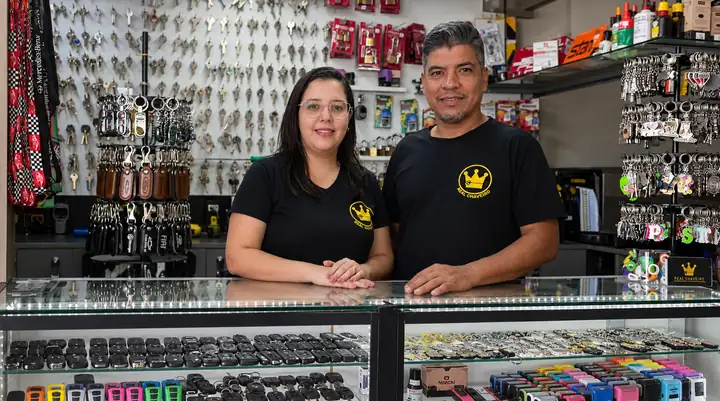 Thatiany e Cleber atrás do balcão de vidro do Real Chaveiro, com a parede de chaves ao fundo