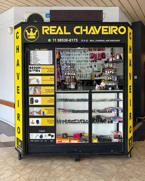 O quiosque do Real Chaveiro dentro do Carrefour Aricanduva: letreiro amarelo com o nome e o telefone, painel de serviços e vitrine de chaves